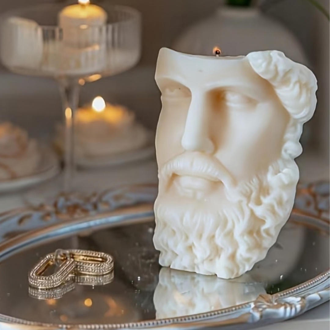 Candle shaped like a classical bust on a reflective surface with candles and flowers in the background.